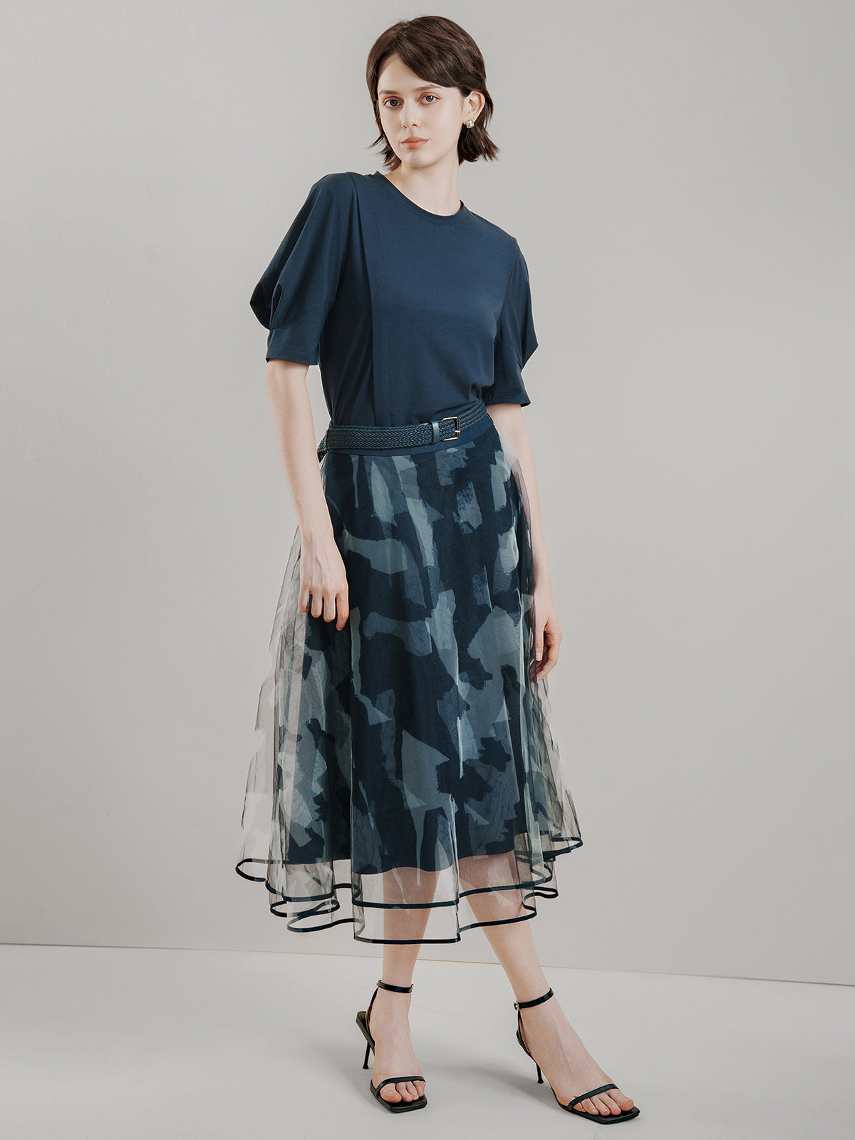 Dark blue printed midi skirt with layered tulle overlay, abstract camouflage pattern, high-waisted A-line silhouette, and matching belt, creating a stylish and edgy look for modern casual wear